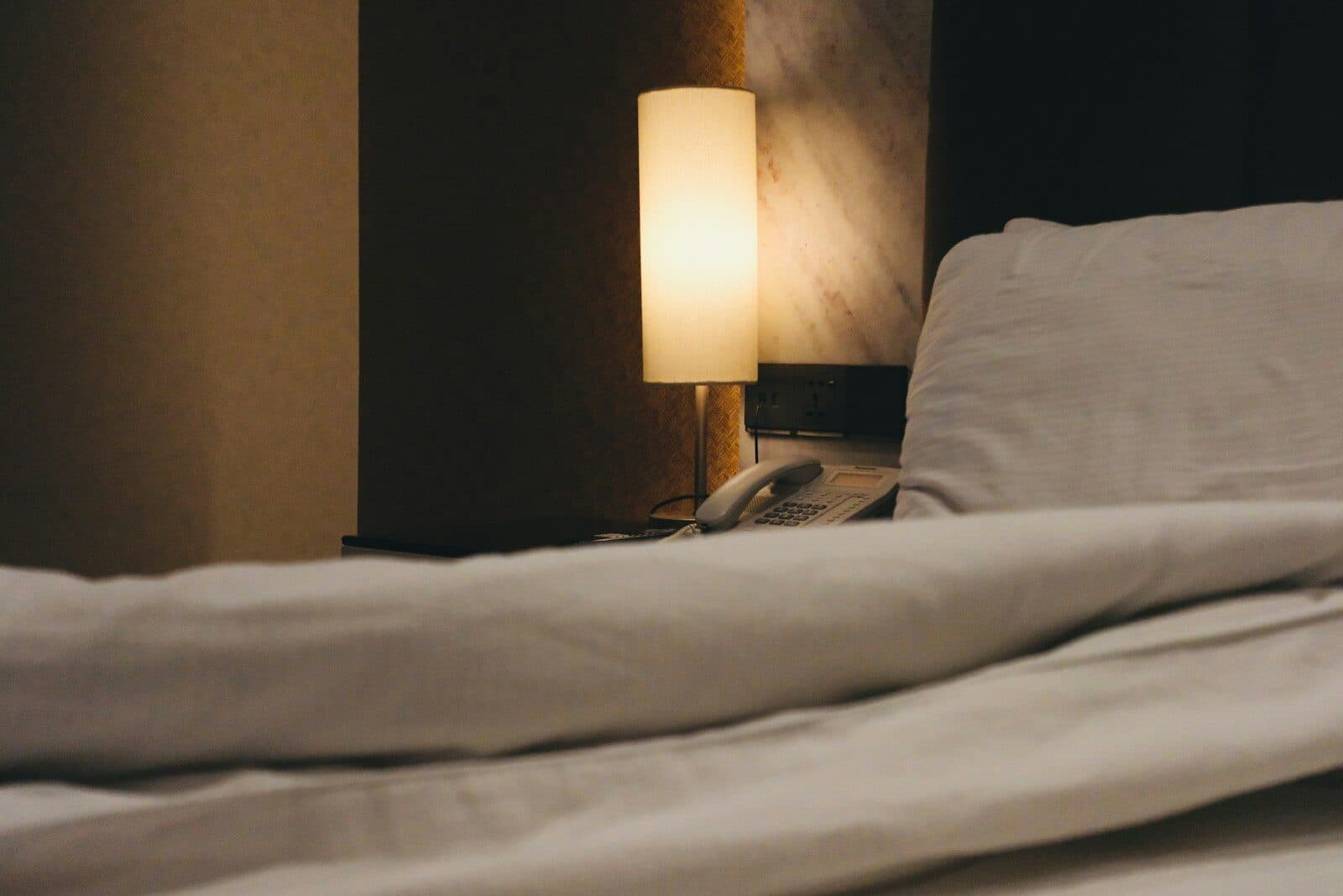 A dimly lit bedside lamp illuminates a rumpled white duvet